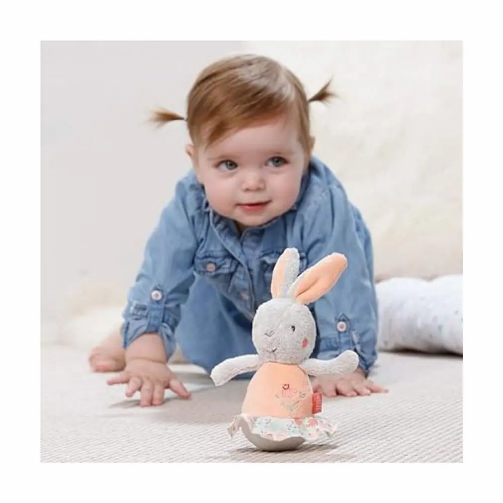 Baby krabbelt auf dem Boden auf ein graues Stofftier-Hase mit rosa Bauch und Ohren zu. Marke: Fehn.