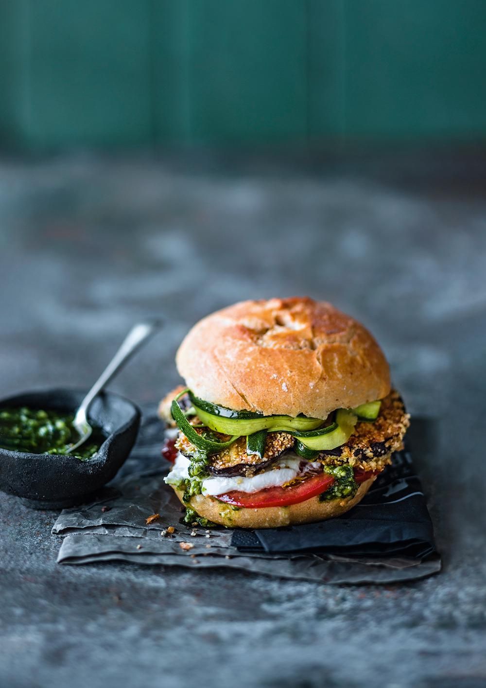 Burger mit Gemüse, Tomate, Pesto. Brötchen, Gurkenscheiben, gebratene Gemüse. Schwarze Schale mit Pesto. Auf schwarzem Papier.