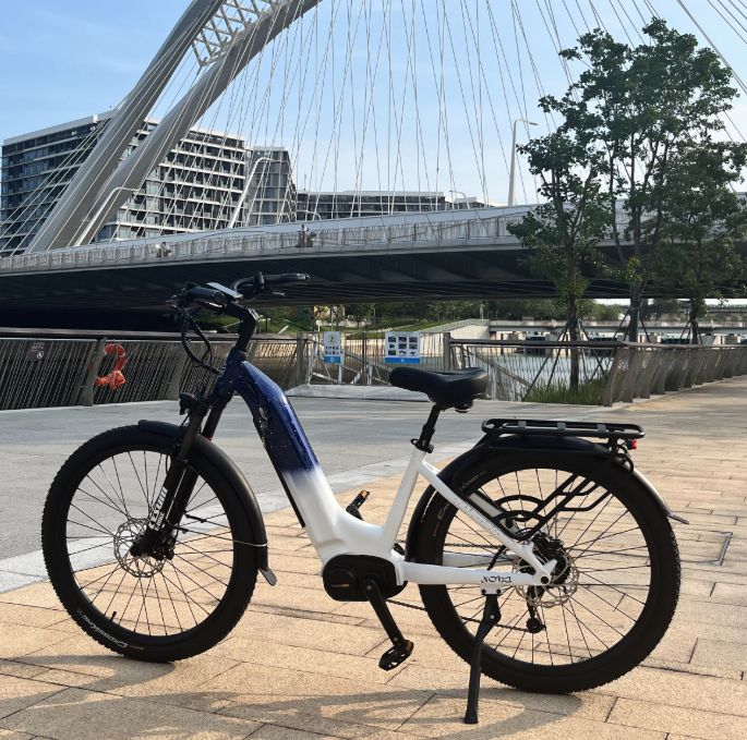 E-Bike mit Mittelmotor, weiß-blau. Brücke im Hintergrund. Schwarze Reifen, Gepäckträger, Sattel.