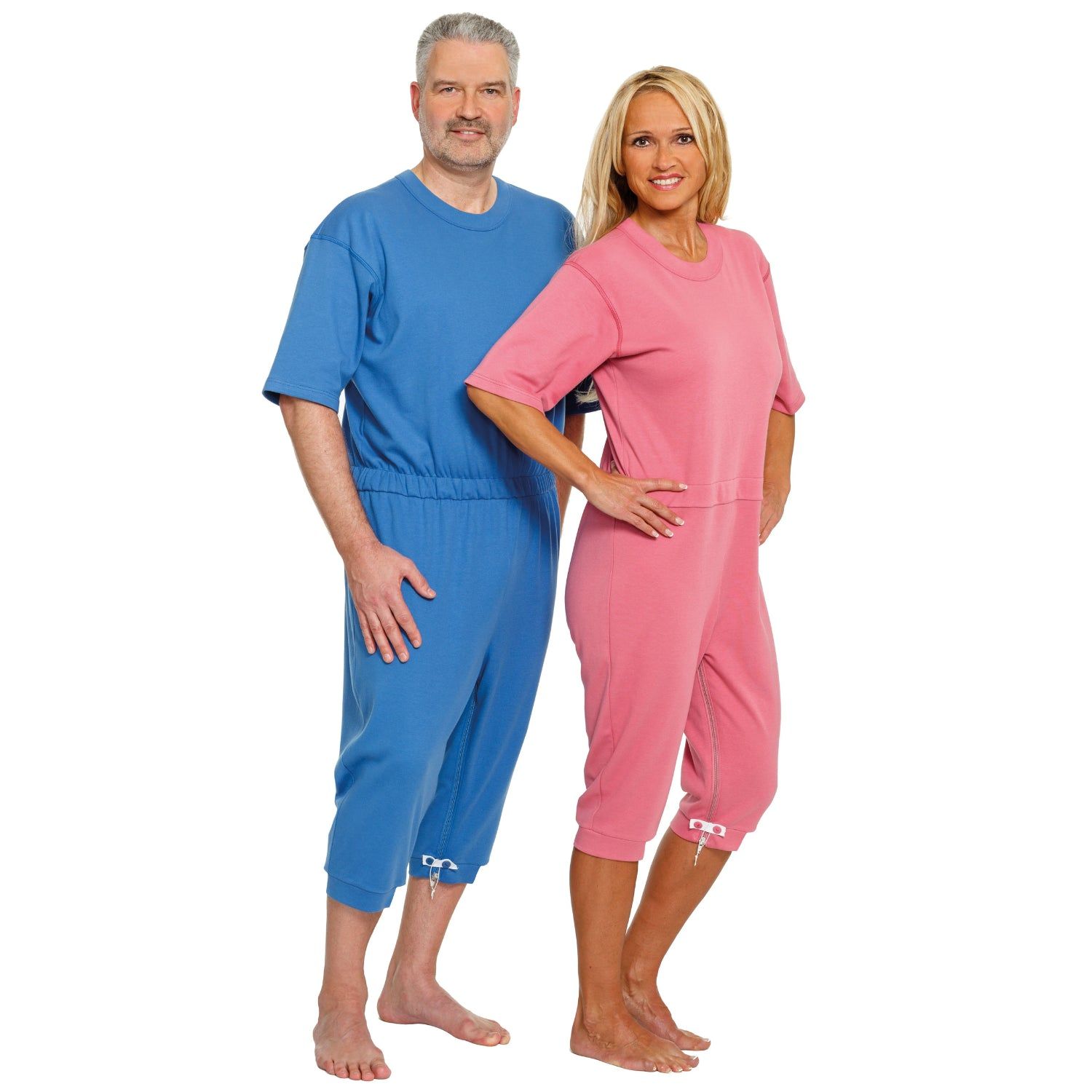 Zwei Personen tragen Pflegeoveralls. Ein Mann in blau, eine Frau in rosa. Beide Overalls haben kurze Ärmel und Hosen.