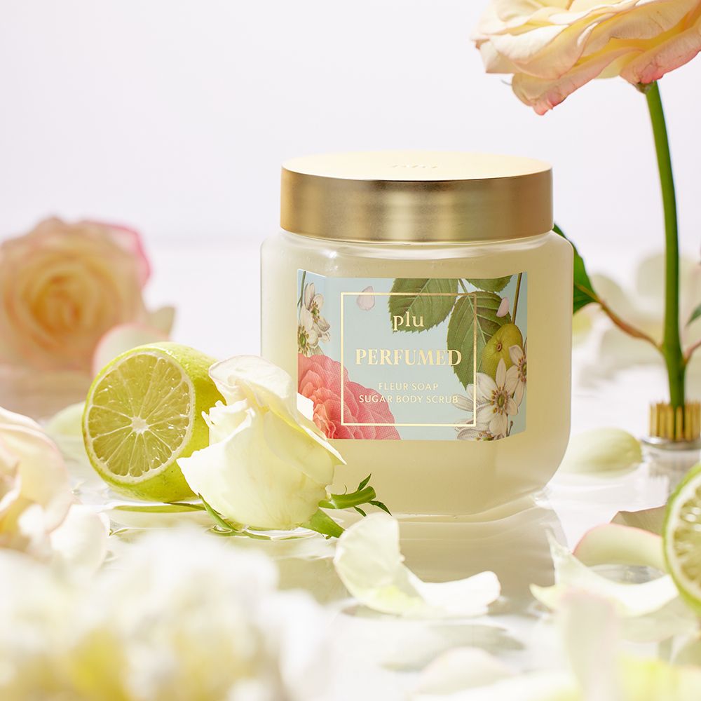Glasbehälter mit goldfarbenem Deckel und Etikett. Umgeben von Blumen und Limetten. Aufschrift: PERFUMED FLEUR SOAP SUGAR BODY SCRUB.