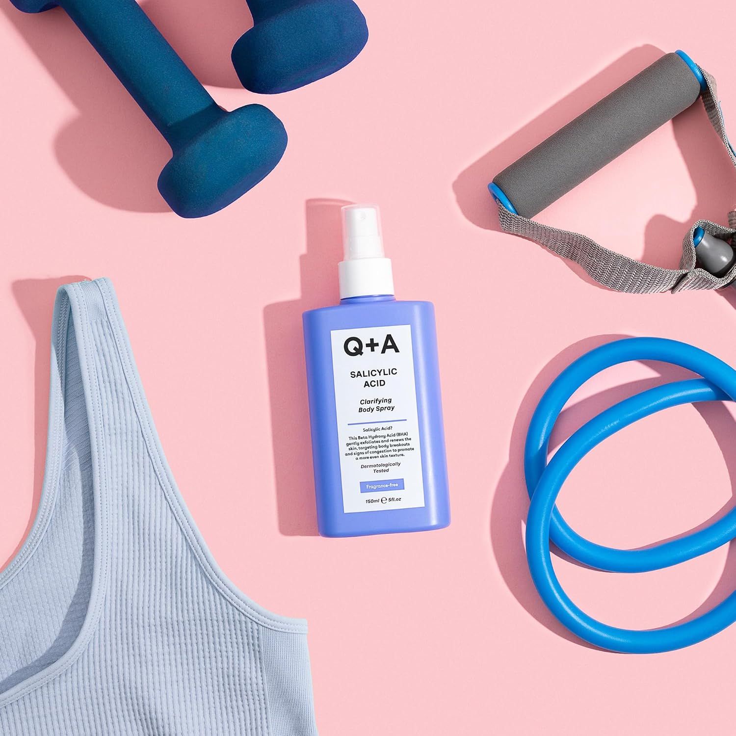 Q+A Salicylic Acid Clarifying Body Spray mit Sportgeräten auf rosa Untergrund.