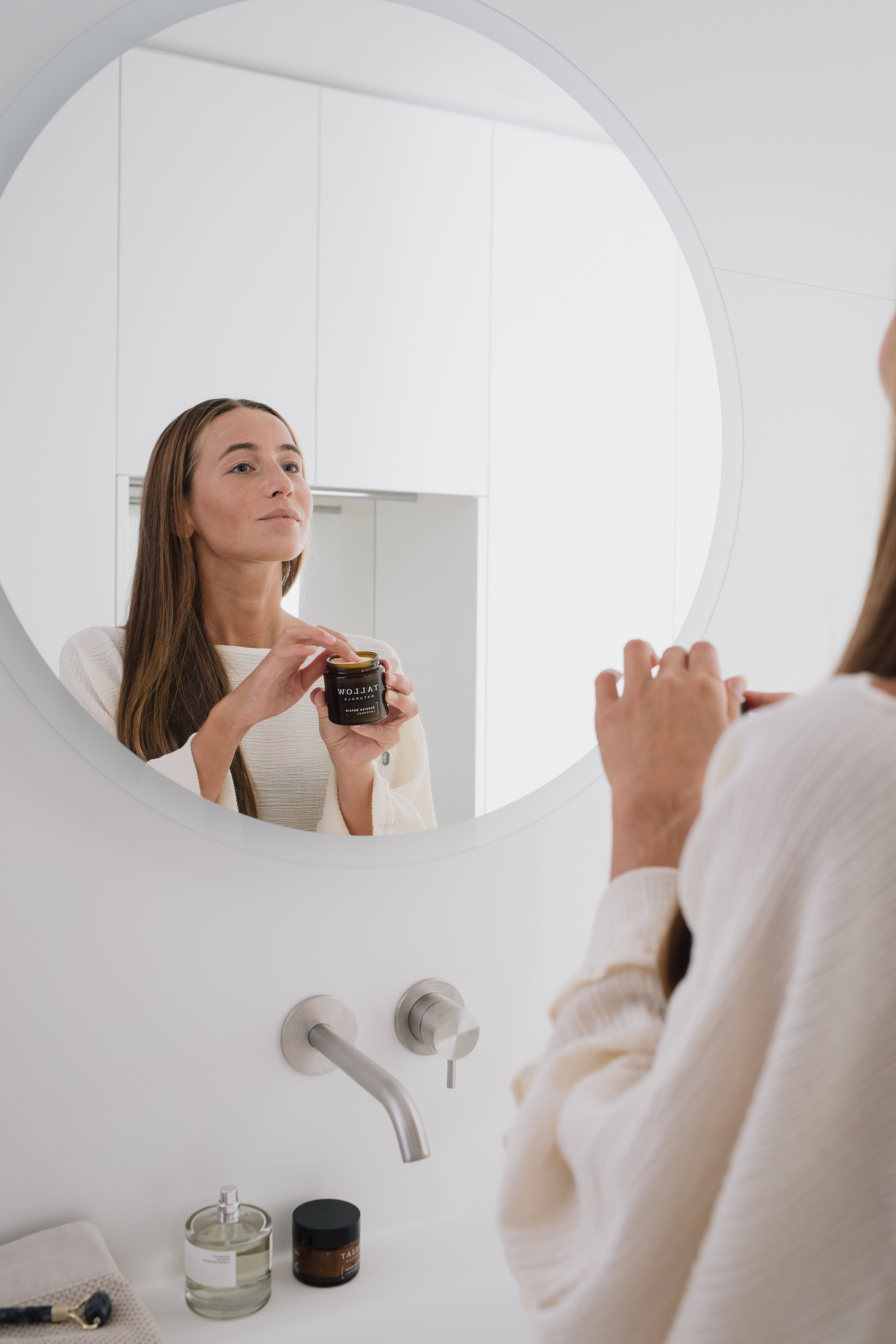 Frau betrachtet sich im Spiegel. Sie hält eine braune Dose mit schwarzem Deckel. Aufschrift: Tallow Naturals, Barrier Repair Pur.