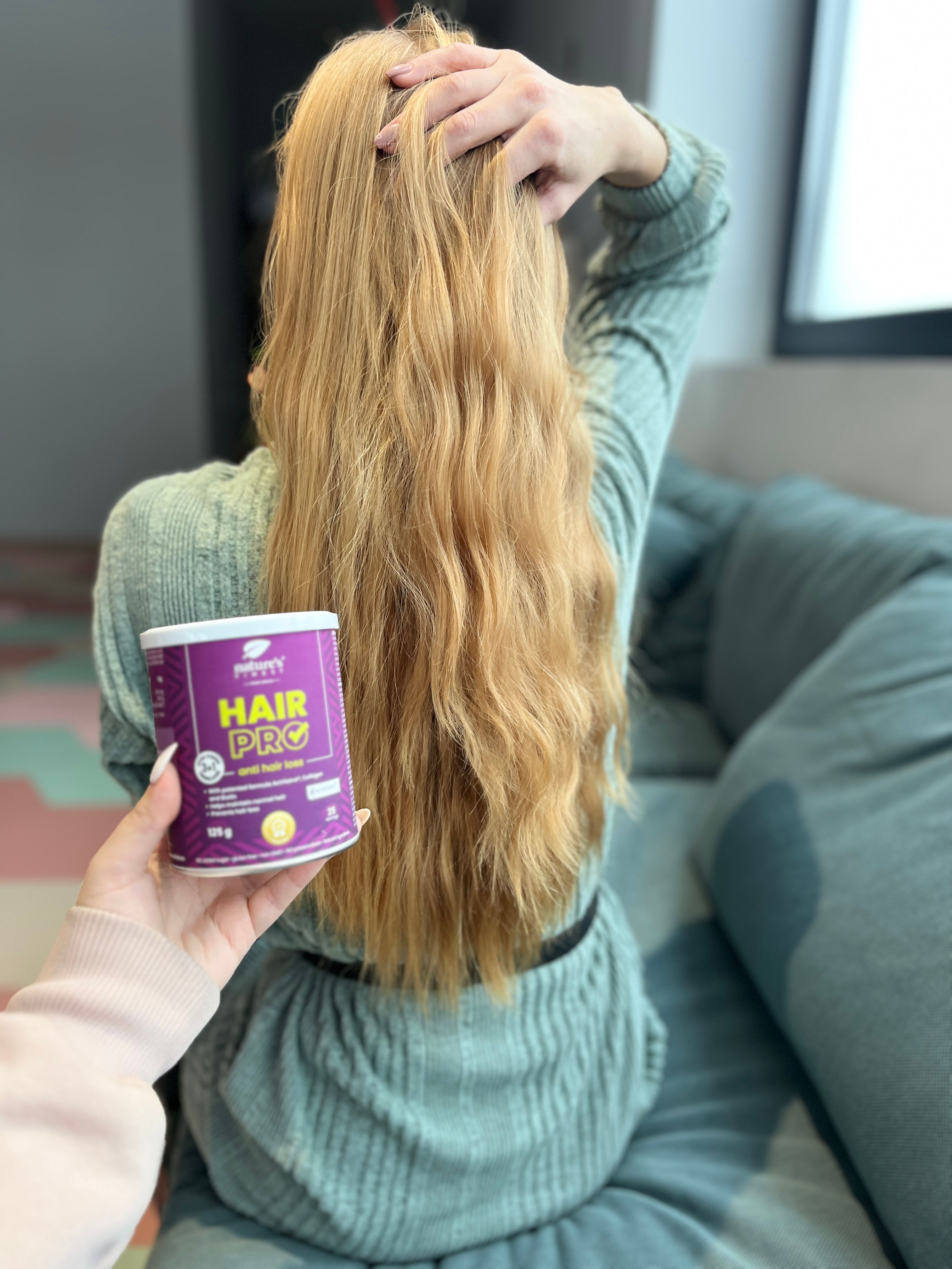 Person hält Dose Nature's Finest Hair Pro. Aufschrift 'Hair Pro' gegen Haarausfall. Lange blonde Haare.