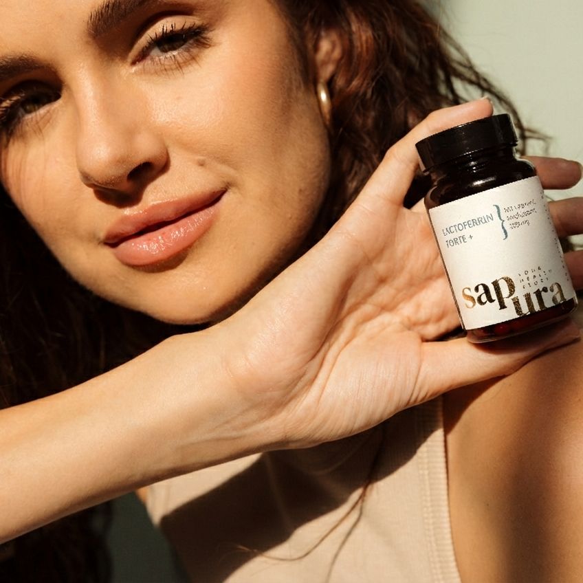 Frau hält eine braune Flasche. Aufschrift: Lactoferrin Forte +, Vitamin C, Sapura. Flasche mit schwarzem Deckel.