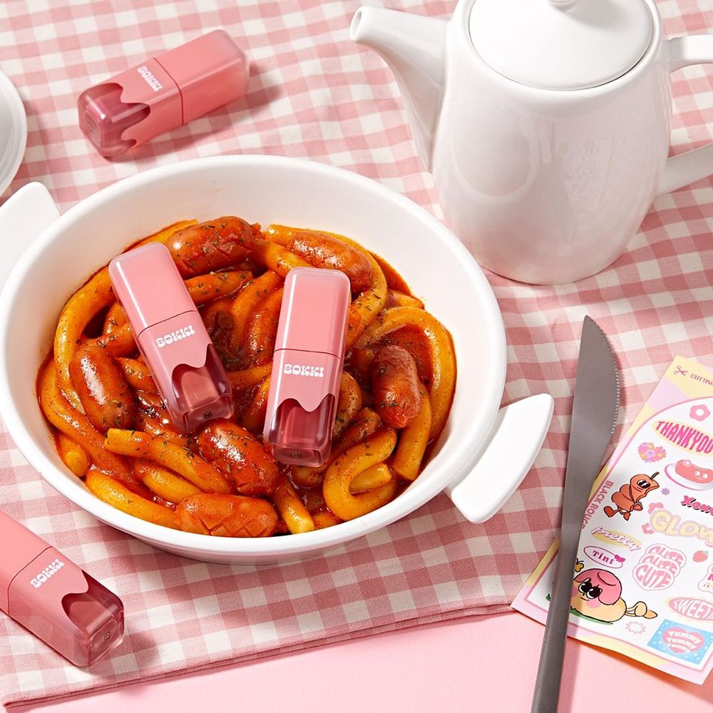 Mehrere rosa Lippenstifte mit "BOKKI"-Schriftzug. Auf einem Tisch mit Essen und einer Teekanne platziert.