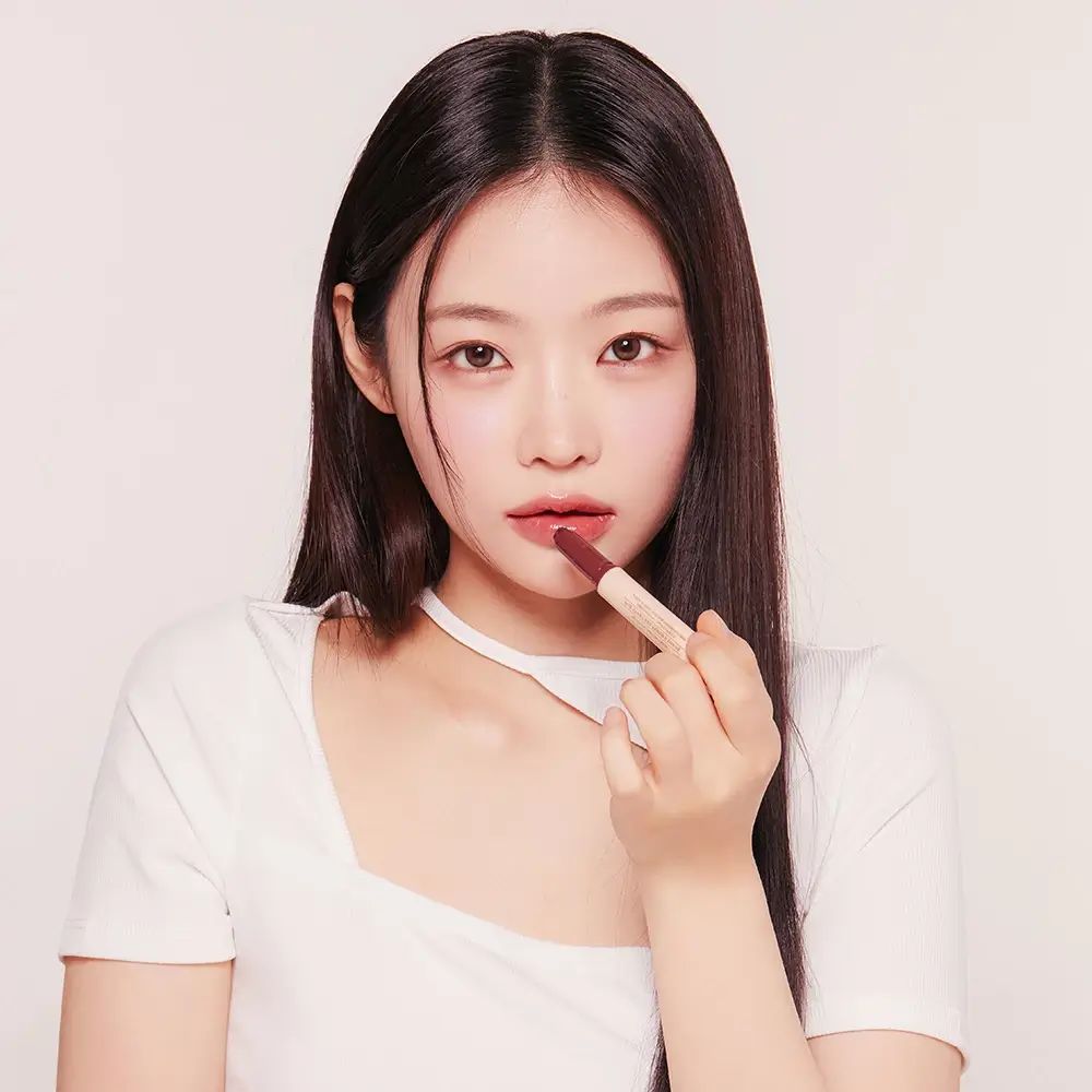 Frau trägt Lipgloss auf. Sie hält einen weißen Stift mit braunem Lipgloss. Hintergrund: weiß.