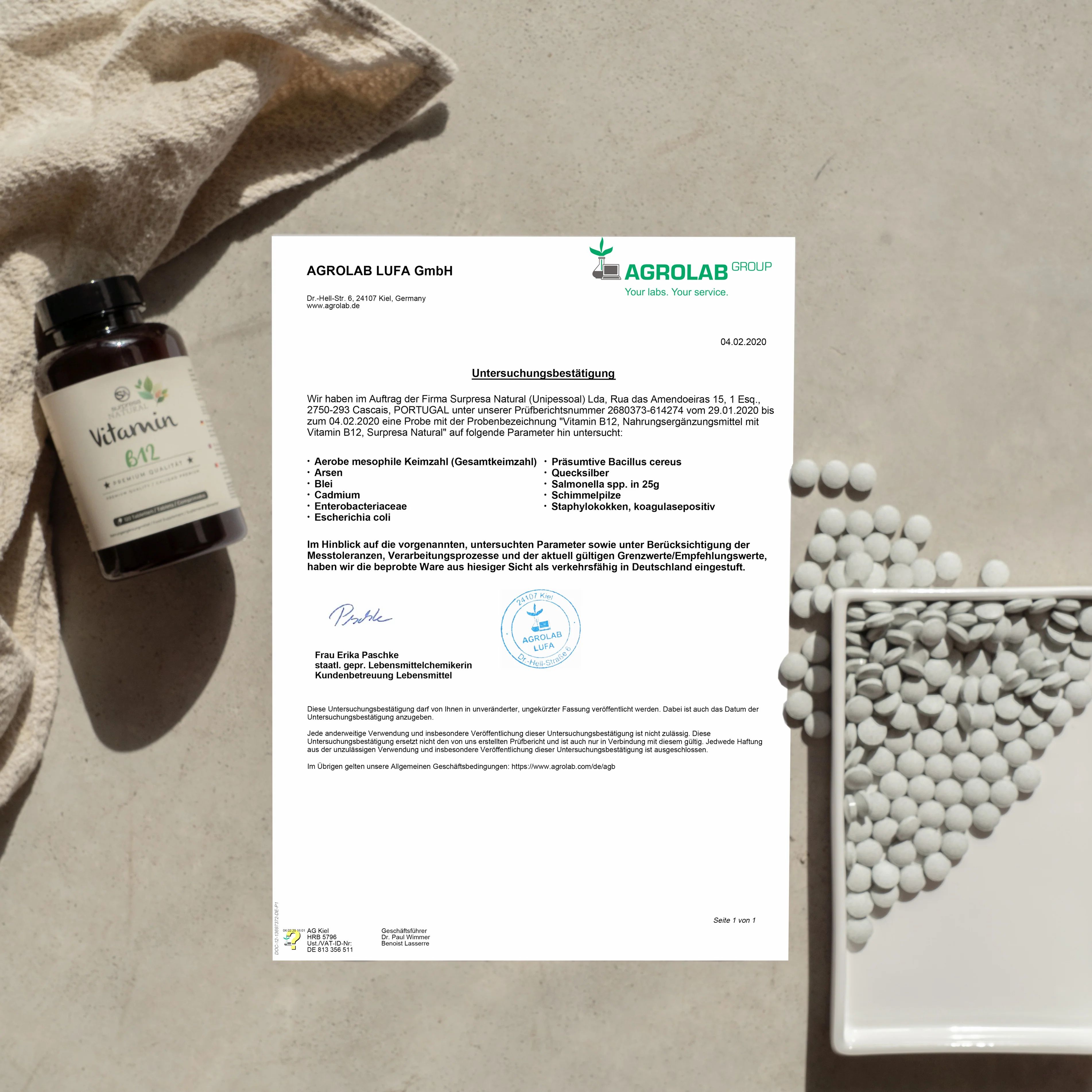 Braune Flasche mit schwarzem Deckel. Etikett mit "Vitamin B". Daneben ein Dokument und Tabletten. Marke: Surpresa Natural.