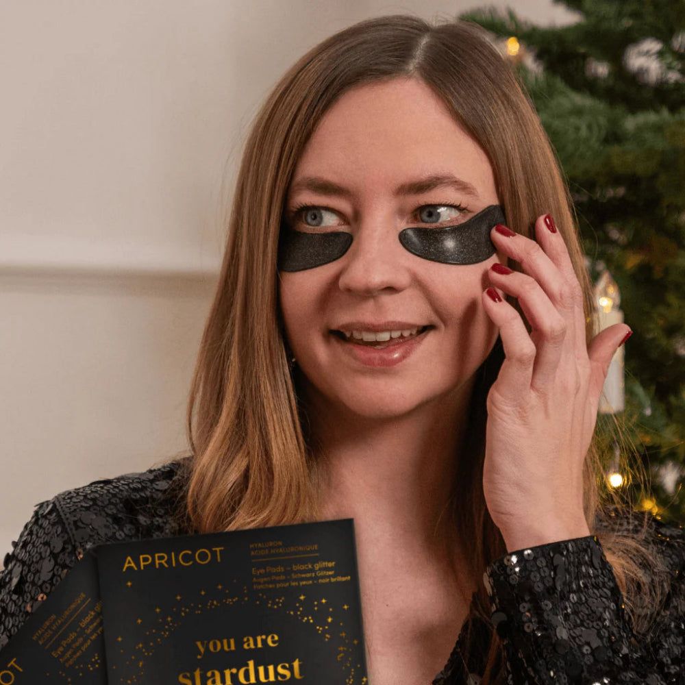 Frau mit schwarzen Glitzer-Augenpads. Hält eine Packung mit APRICOT-Produkt. Aufschrift: "you are stardust".
