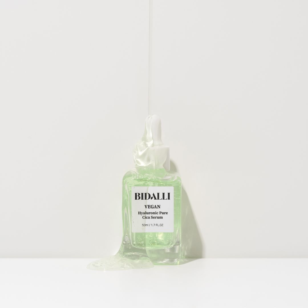 Glasflasche mit grünlicher Flüssigkeit und Pipette. Etikett mit 'Bidalli Vegan Hyaluronic Pure Cica Serum'. Flüssigkeit tropft.