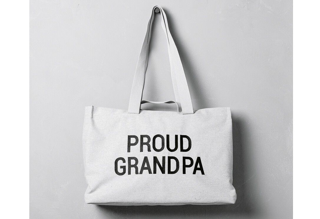 Graue Leinwandtasche mit Tragegriffen. Schwarze Aufschrift "PROUD GRANDPA". Hängt an einer Wand.