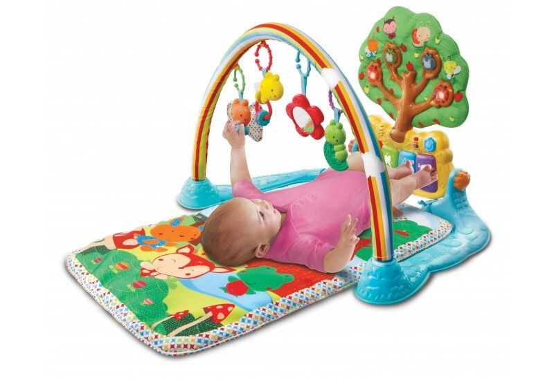 Baby liegt auf Spieldecke. Spielbogen mit Anhängern, Baum mit Tasten und Spielzeug. Bunte Waldmotive auf der Decke.