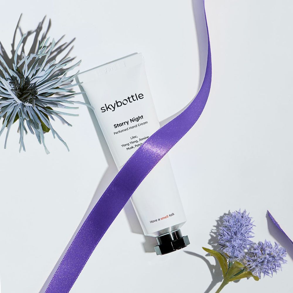 Weiße Tube "skybottle Starry Night Perfumed Hand Cream" mit lila Band und Blumen. Schwarzer Verschluss. Auf hellem Hintergrund.