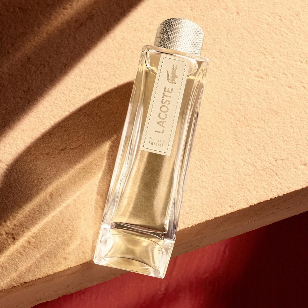 Lacoste Pour Femme EdP Nat. Spray. Flakon auf beiger Oberfläche. Rechteckiger Flakon mit goldfarbenem Verschluss. Logo und Schriftzug.