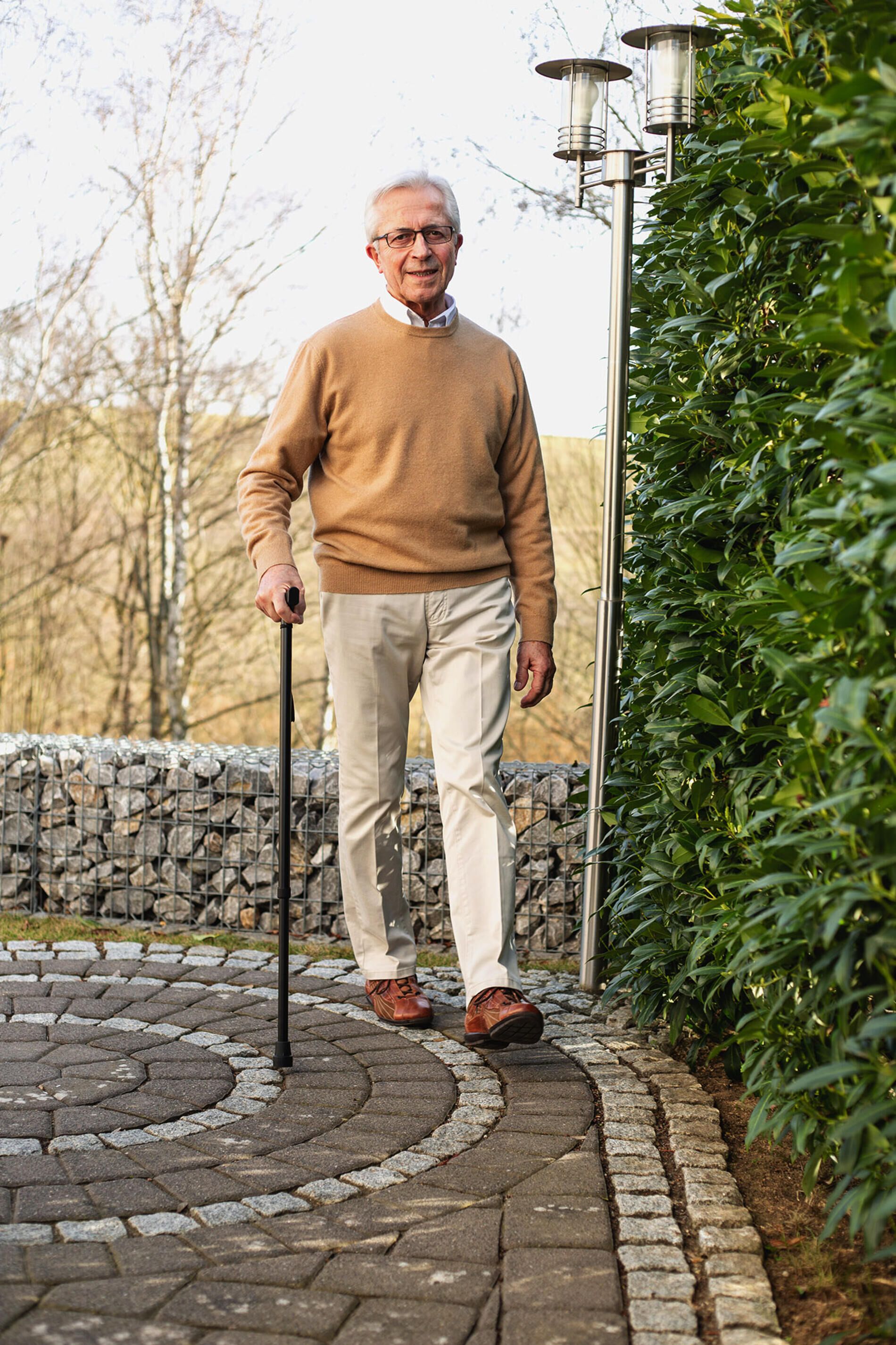 Älterer Mann steht auf einem gepflasterten Weg und stützt sich auf einen schwarzen Gehstock. Er trägt einen beigen Pullover und eine helle Hose.