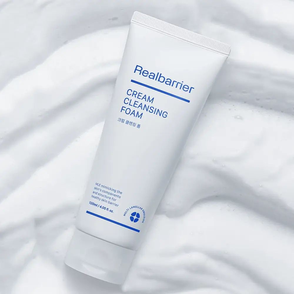 Weiße Tube mit blauem Text. Aufschrift: Realbarrier, CREAM CLEANSING FOAM. Auf Schaum gelegt.