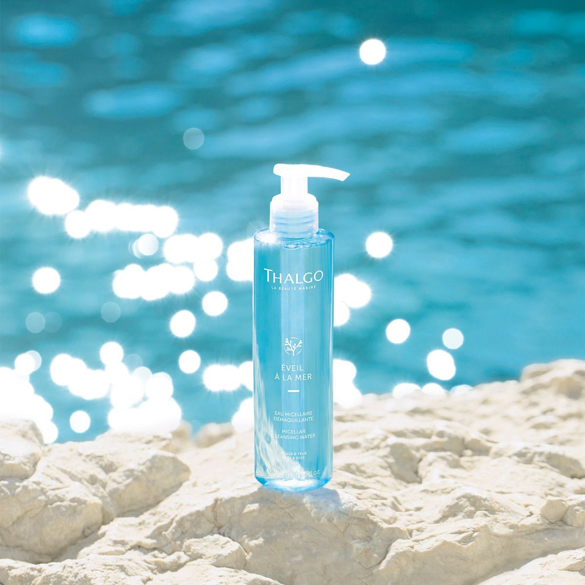Blaues Reinigungswasser in einer Flasche mit weißem Pumpverschluss. Aufschrift: THALGO, EVEIL À LA MER, Micellar Cleansing Water. Vor blauem Wasser.