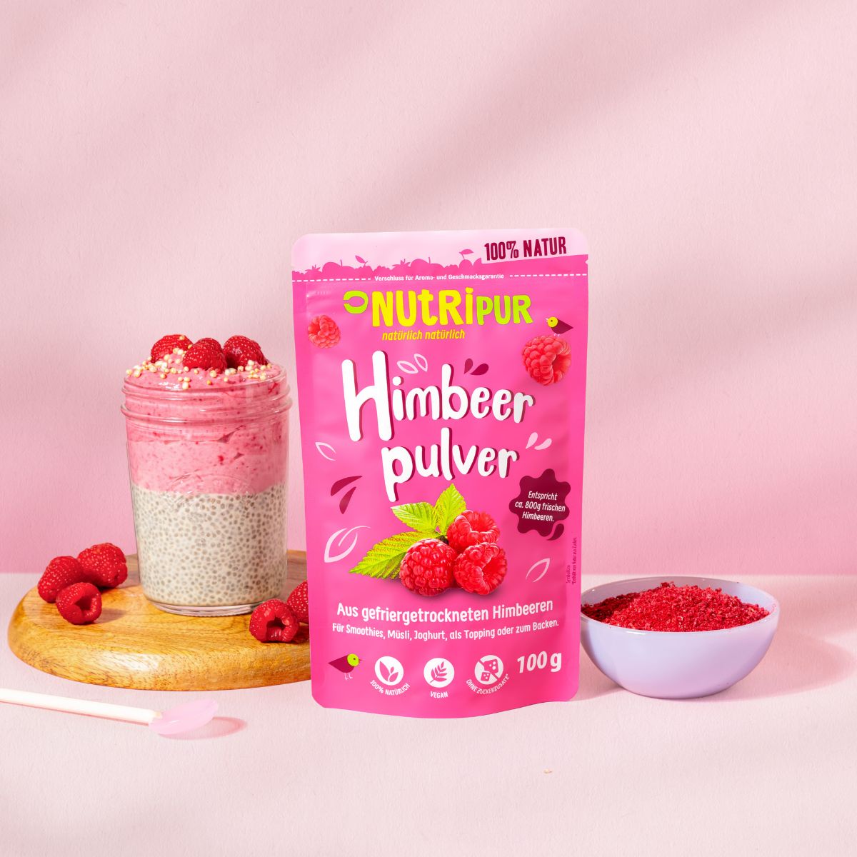Rosa Beutel mit Himbeerpulver. Aufschrift: NutriPur, Himbeerpulver, 100g. Glas mit Chia-Pudding, Himbeeren, Schale mit Pulver.