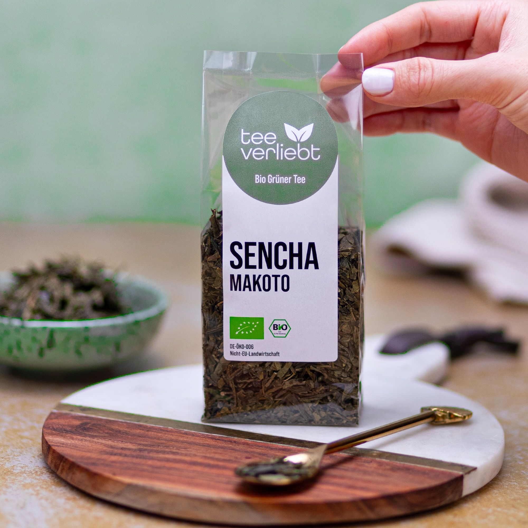 Hand hält Verpackung mit losem Grüntee. Auf Holzbrett mit Löffel. Aufdruck: "tee verliebt", "Sencha Makoto", Bio-Siegel.