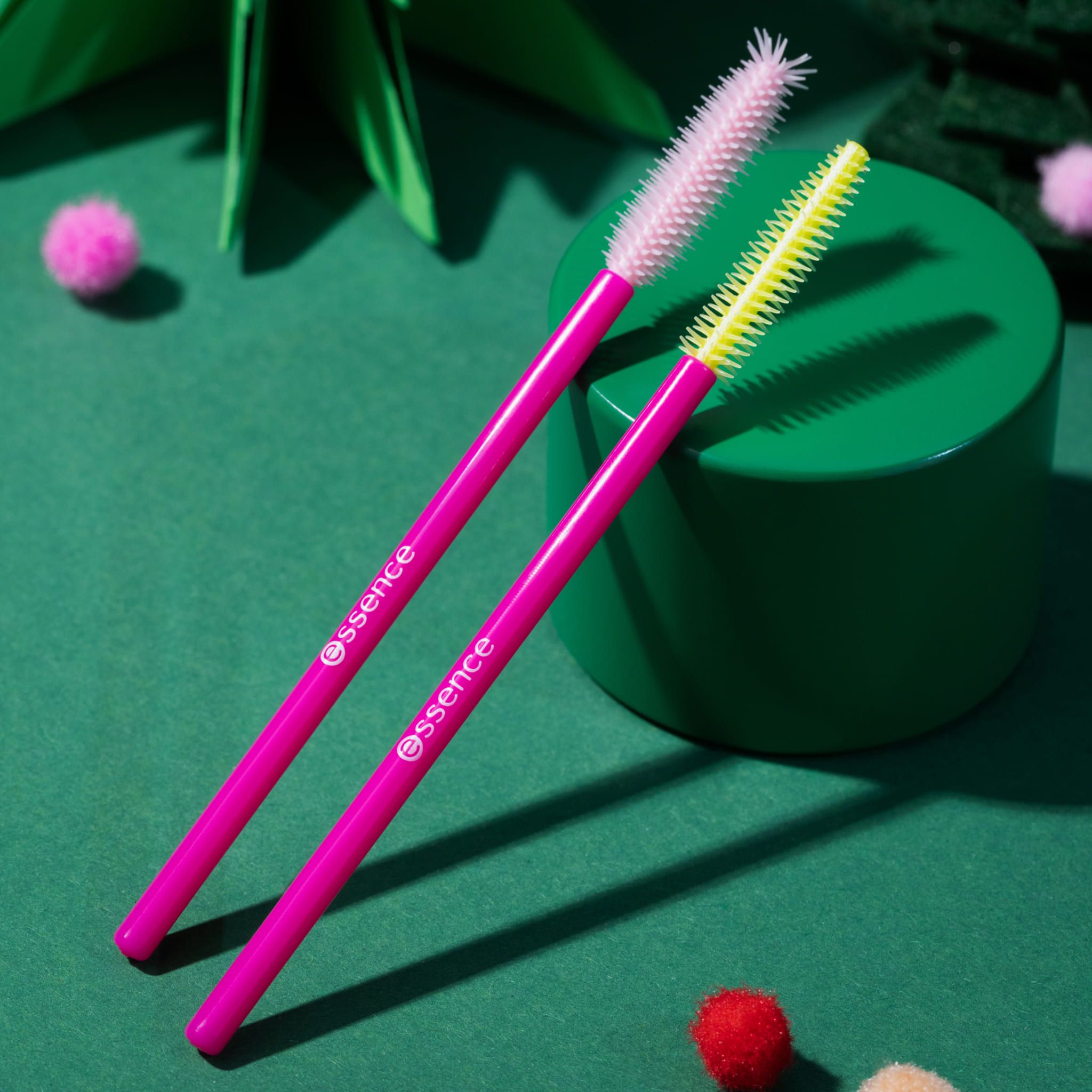 Zwei pinkfarbene Wimpernbürsten mit gelben Borsten. Markenlogo auf dem Griff. Auf grünem Hintergrund.