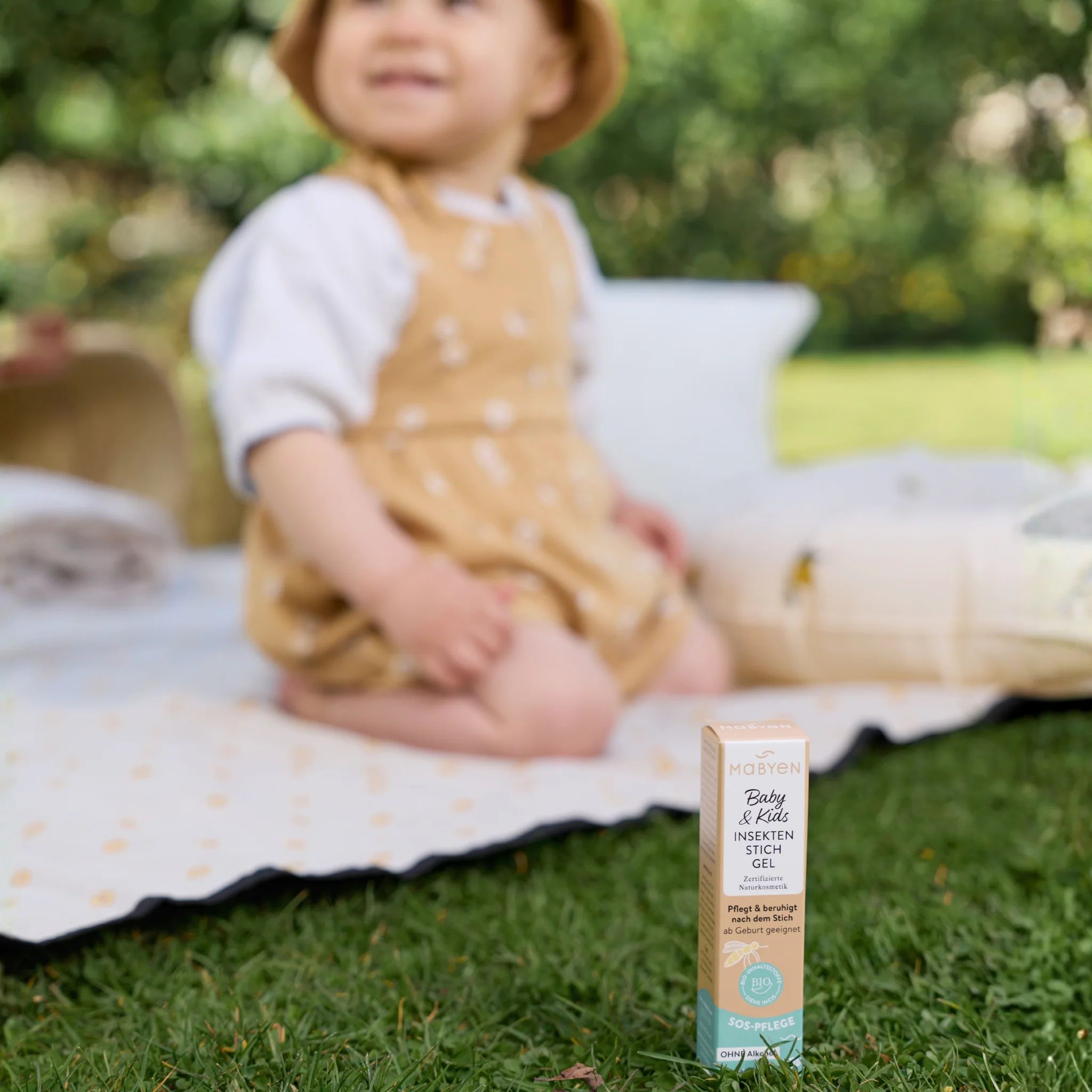 Eine MABYEN Insektenstich Gel Packung steht auf Gras, ein Baby sitzt im Hintergrund.