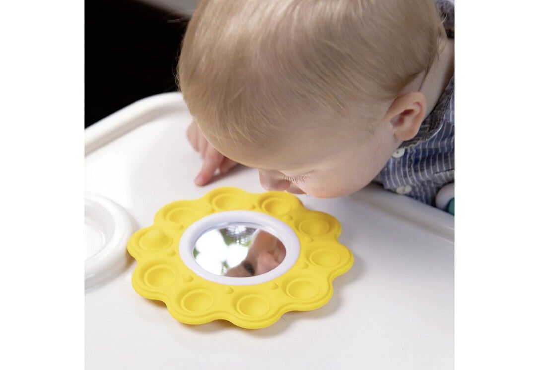 Ein Baby betrachtet ein gelbes, blütenförmiges Spielzeug mit einem Spiegel in der Mitte. Das Spielzeug liegt auf einem weißen Tablett.
