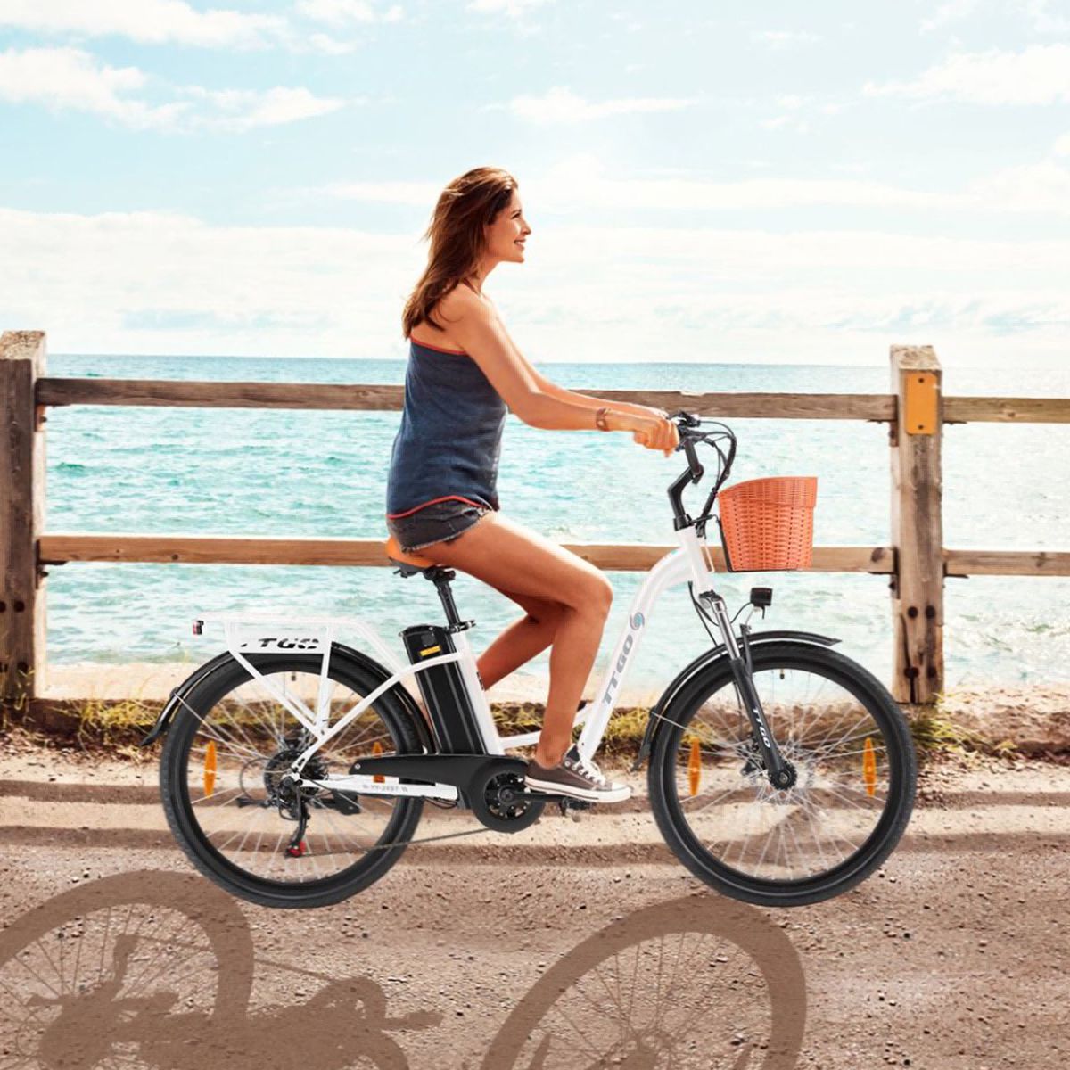 Frau fährt weißes Elektrofahrrad mit Korb. Brauner Sattel und Lenkergriffe. TTGO-Logo. Hintergrund: Meer.