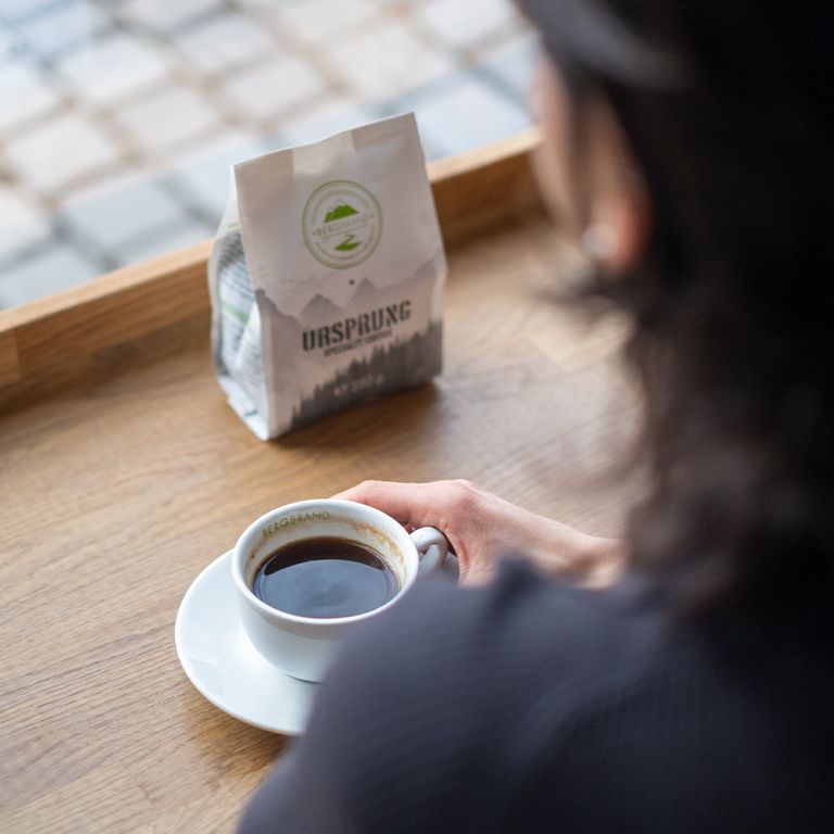 Kaffeetasse und Kaffeebohnen-Packung. Packung mit Logo und Text Ursprung. Auf Holztisch am Fenster.
