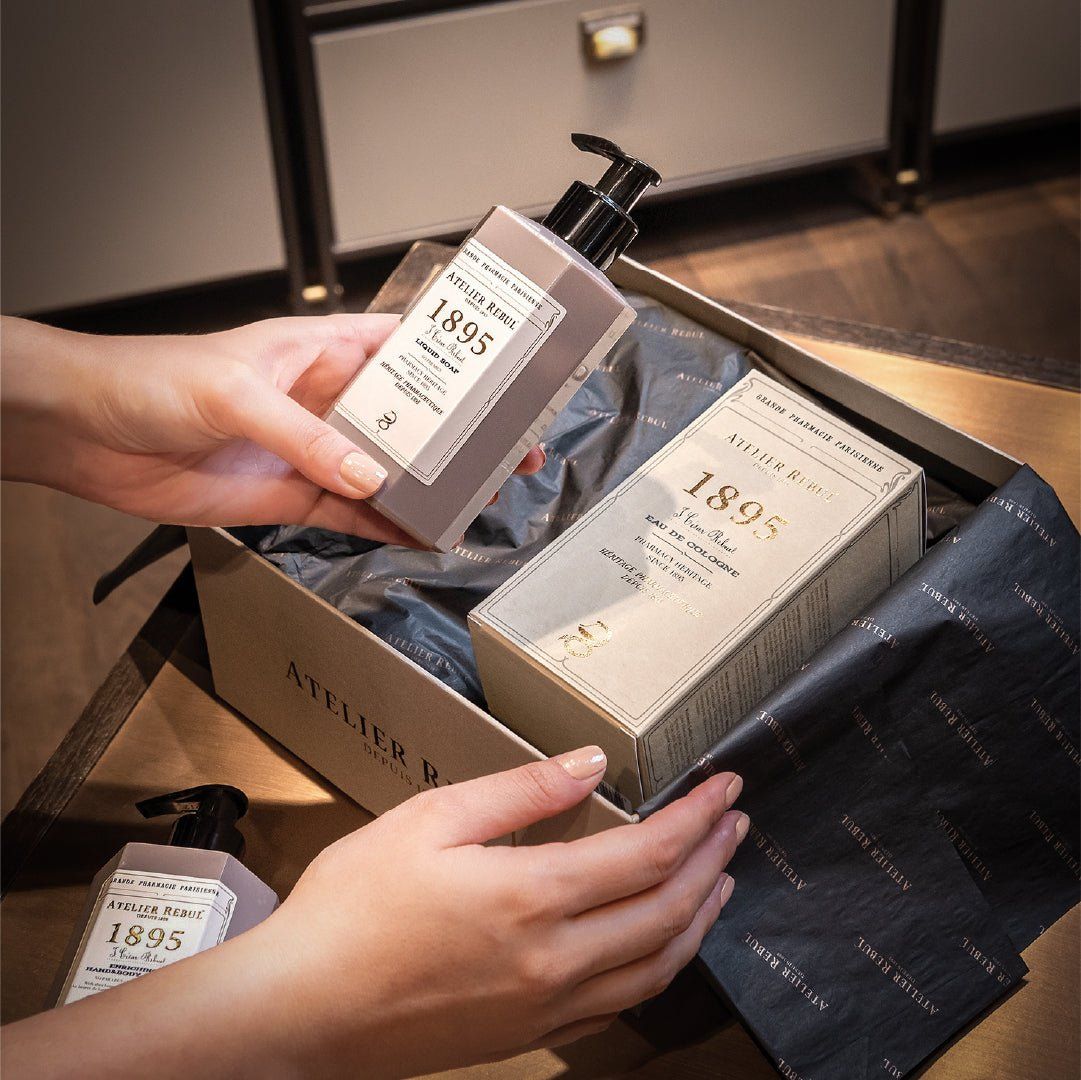Hände halten eine Flasche und eine Schachtel. Aufschrift: Atelier Rebul 1895. In einer Geschenkbox.
