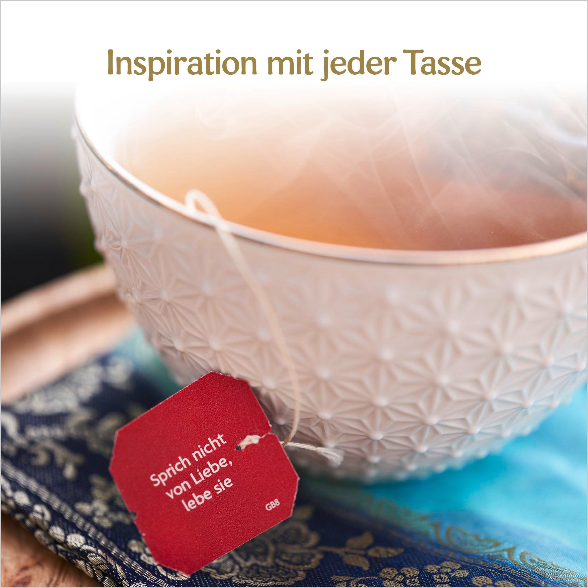 Nahaufnahme einer Tasse Tee mit Teebeutel. Roter Anhänger mit Text. Text: Inspiration mit jeder Tasse. Auf blauem Stoff.