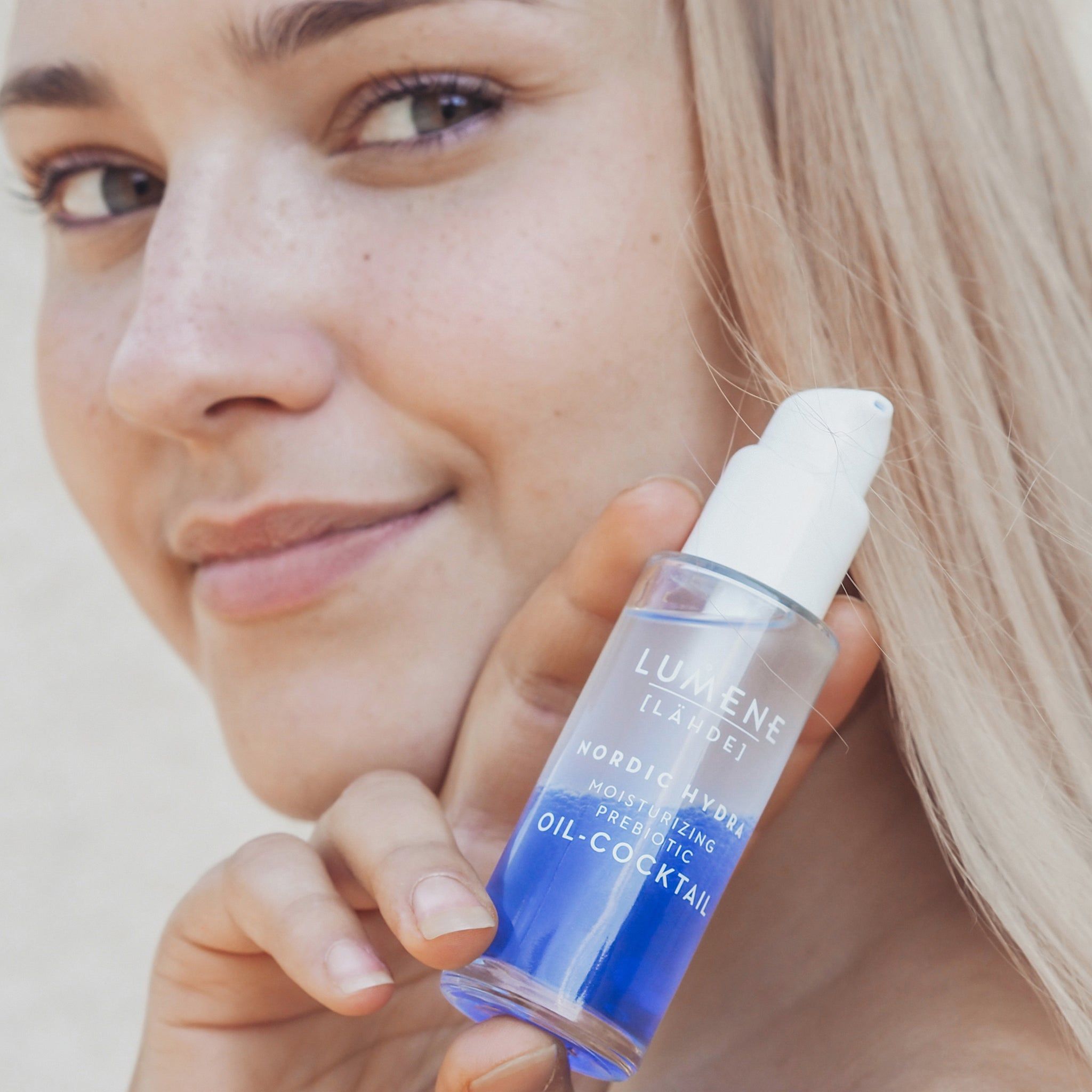 Frau hält einen Flakon. Flakon mit weißem Pumpkopf. Blaue und transparente Flüssigkeit sichtbar. Produktname und Marke.
