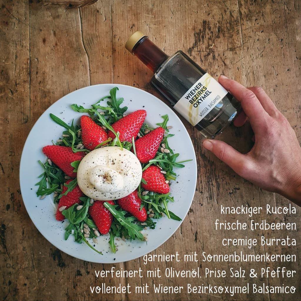 Eine Flasche mit klarer Flüssigkeit, gehalten von einer Hand. Auf einem Teller: Salat, Erdbeeren und Burrata.