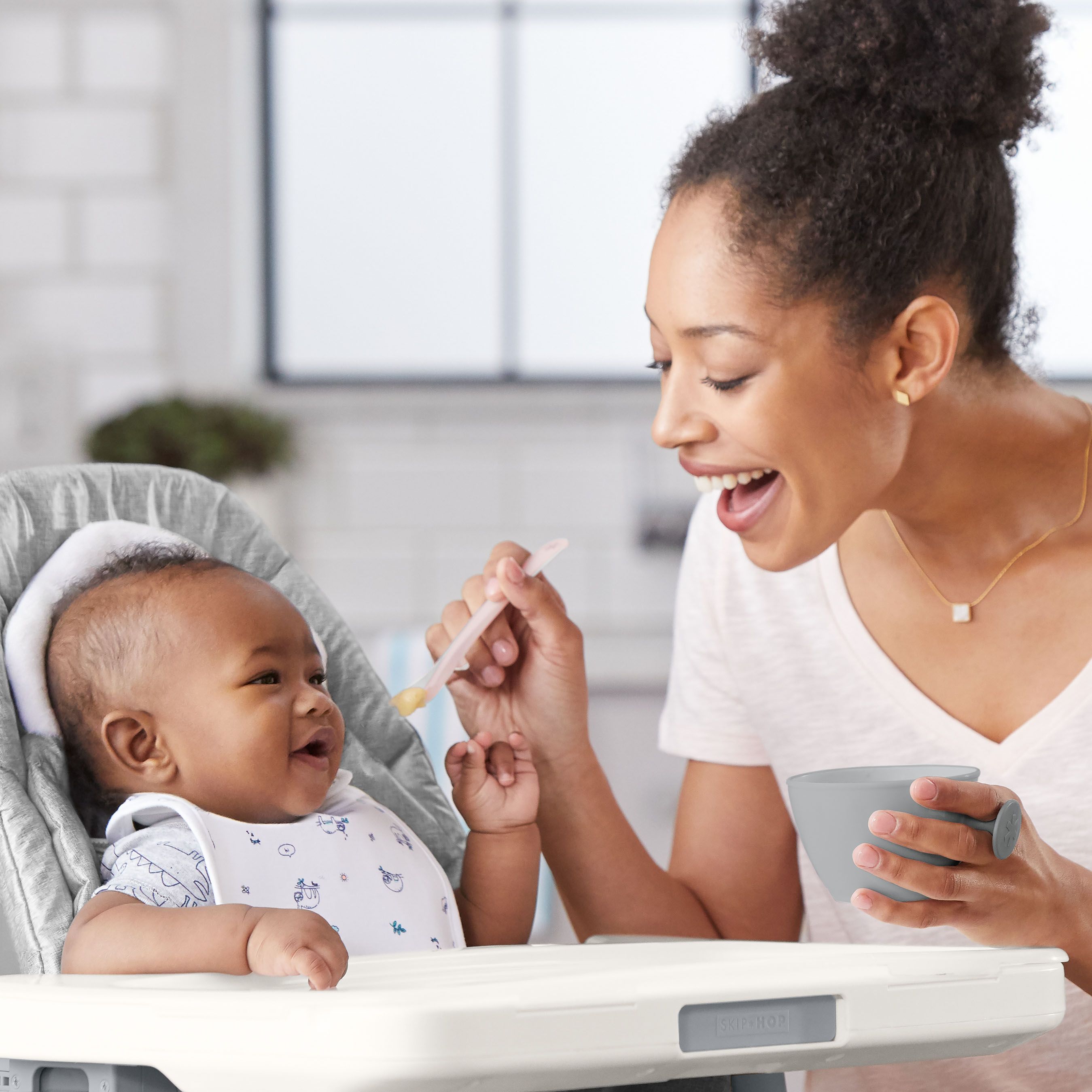 Eine Frau füttert ein Baby mit einem Löffel. Das Baby sitzt in einem Hochstuhl. Die Frau hält eine Schüssel.