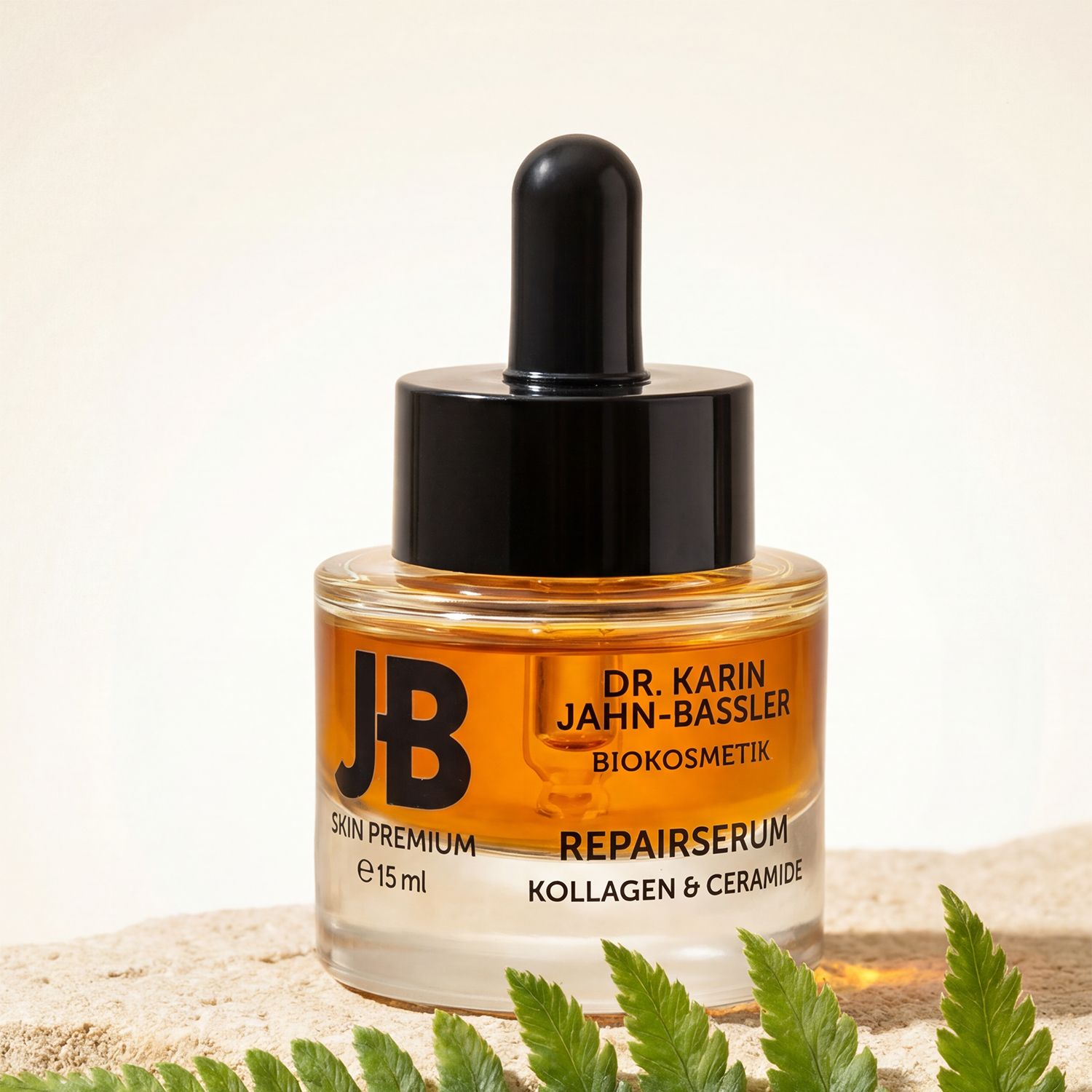 Serumflasche mit schwarzem Deckel und Pipette auf Sand. Aufschrift: JB Skin Premium, Dr. Karin Jahn-Bassler, Repaierserum, Kollagen & Ceramide.