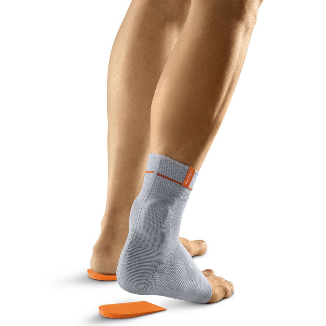 Graue Achillessehnen-Bandage am Fuß. Orangefarbene Akzente. Fußsohle mit orangefarbener Einlage. Person steht auf weißem Hintergrund.