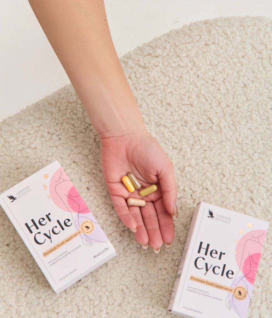 Zwei "Her Cycle" Verpackungen neben einer Hand mit Kapseln. Logo und Illustration einer Frau. Marke: Carnium Botanicals.
