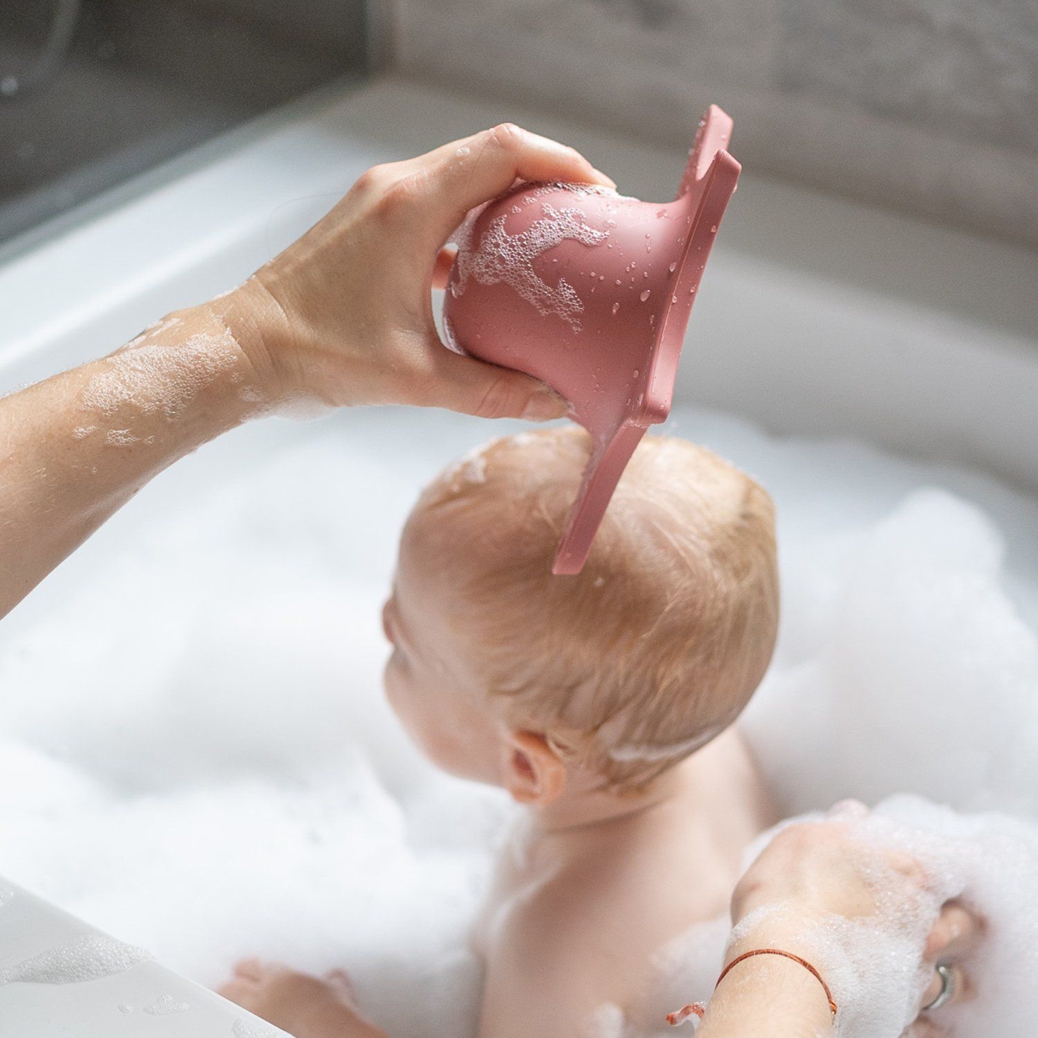 Person hält rosa Duschhilfe über einem Baby in einer Badewanne. Das Baby ist von Schaum umgeben.