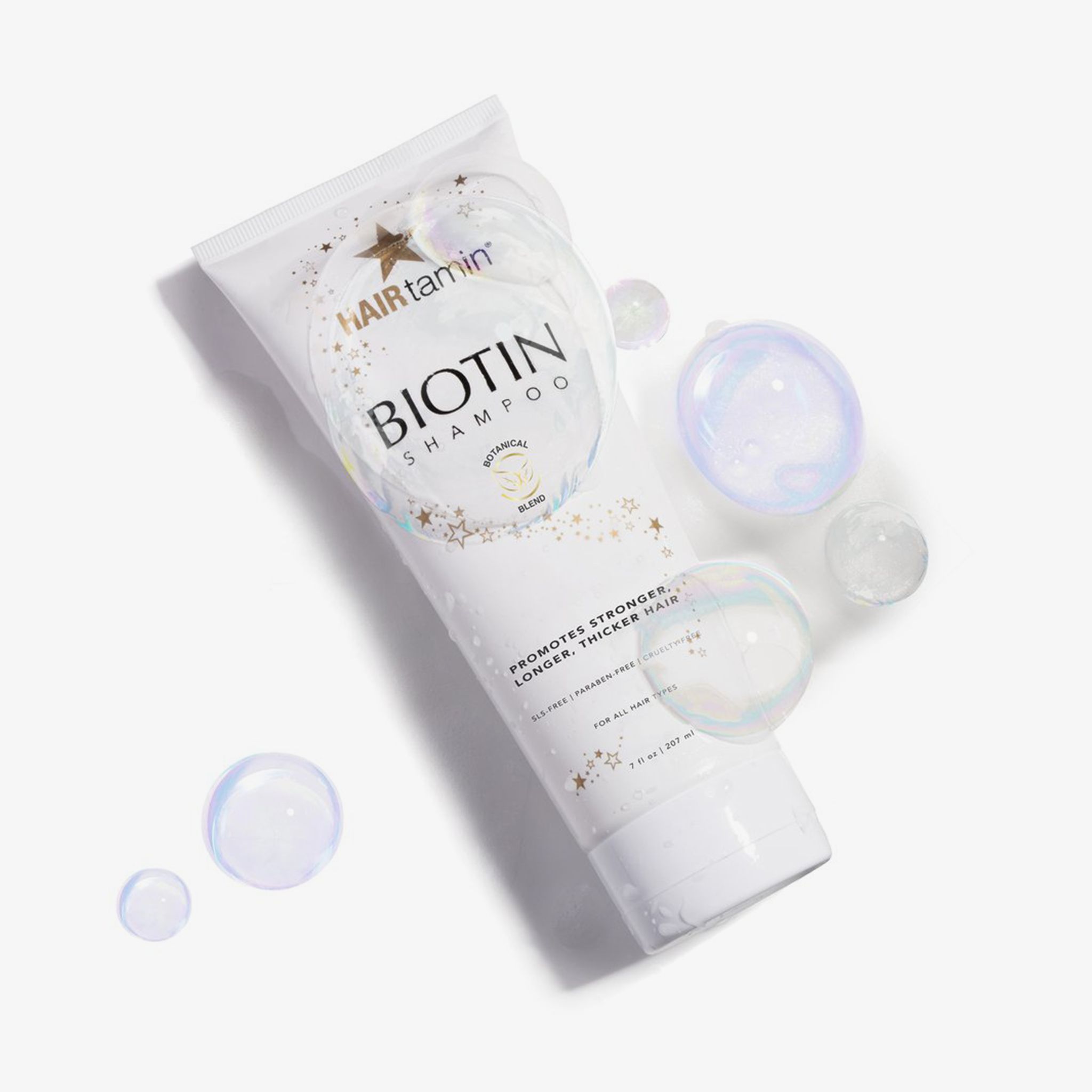Weiße Tube "HAIRtamin Biotin Shampoo" mit Seifenblasen. Sterne und Schriftzug "Botanical Blend". Text: "Supports stronger, longer, thicker hair".