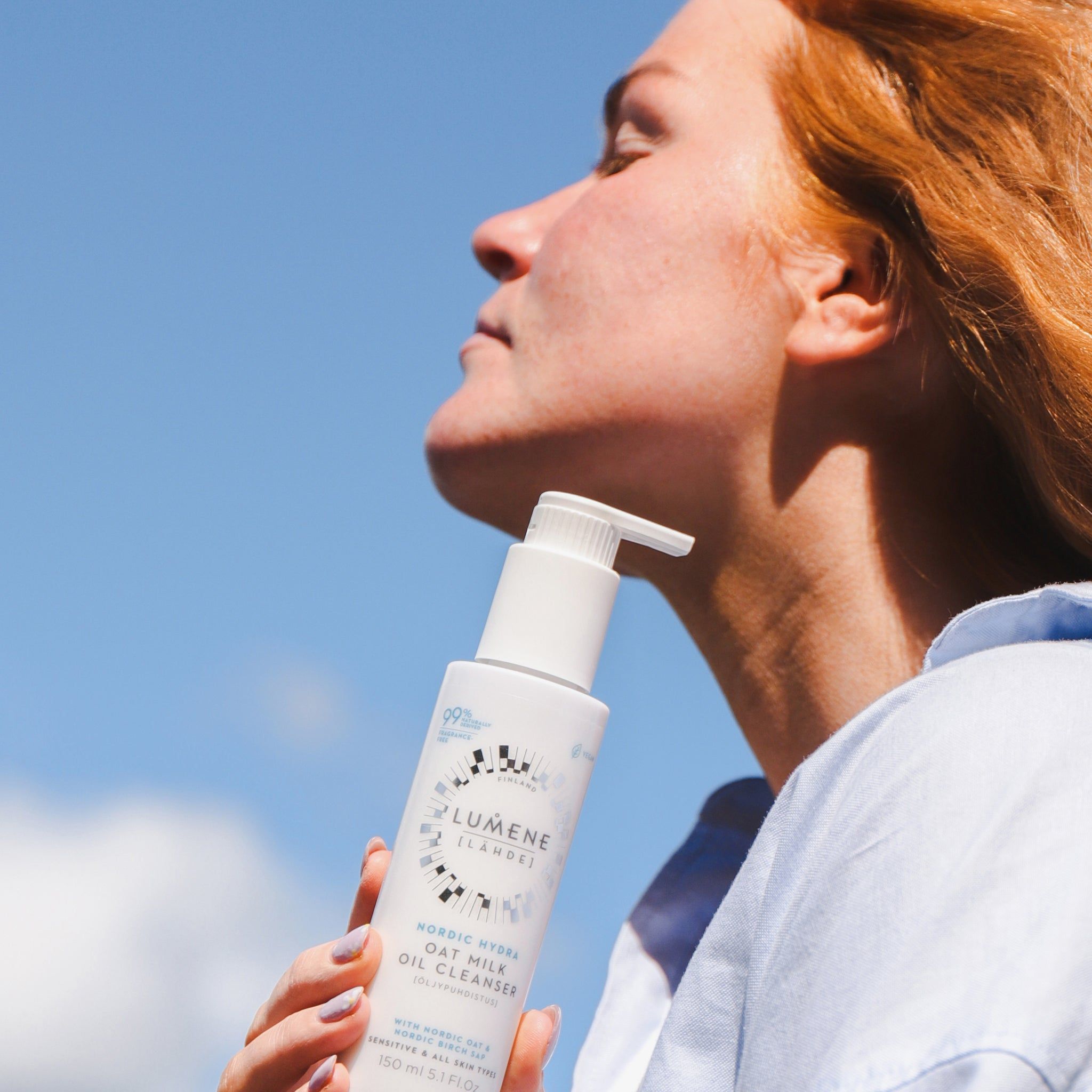 Frau hält eine weiße Flasche mit Pumpe. Aufschrift: Lumene Nordic Hydra Oat Milk Oil Cleanser. 99% natürlich. Blauer Himmel.