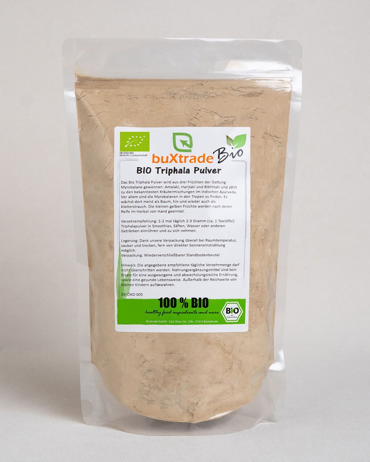 Bio Triphala Pulver in einem Standbodenbeutel mit Etikett. Daneben eine kleine Schale mit Pulver und ein Glas Milch.