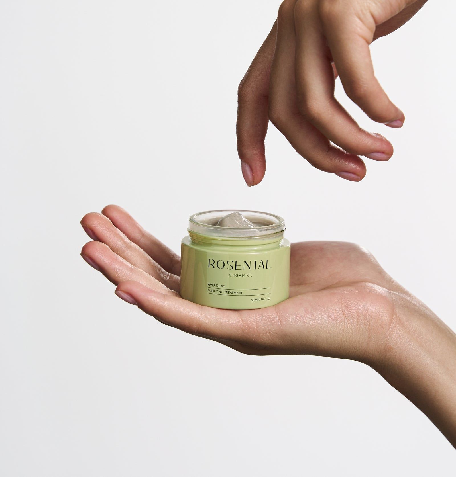 Hände halten ein grünes Rosental-Glas mit Avo Clay Mask. Eine weitere Hand schwebt darüber. Helle Hintergrund.