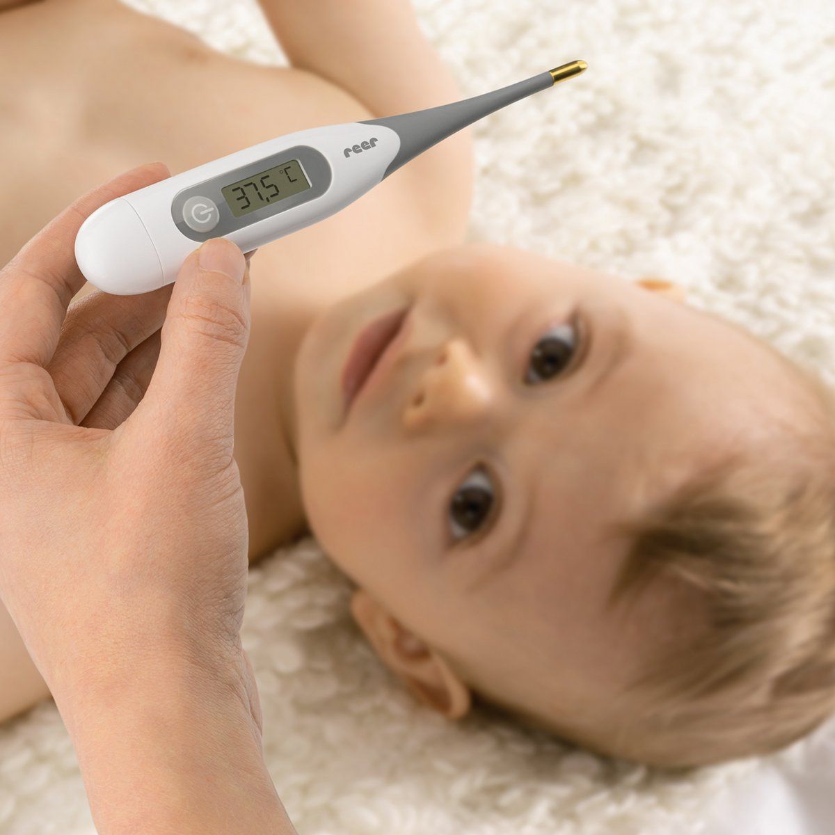 Ein Thermometer wird an einem Baby gemessen. Das Thermometer zeigt 37,5°C an. Marke: reer.
