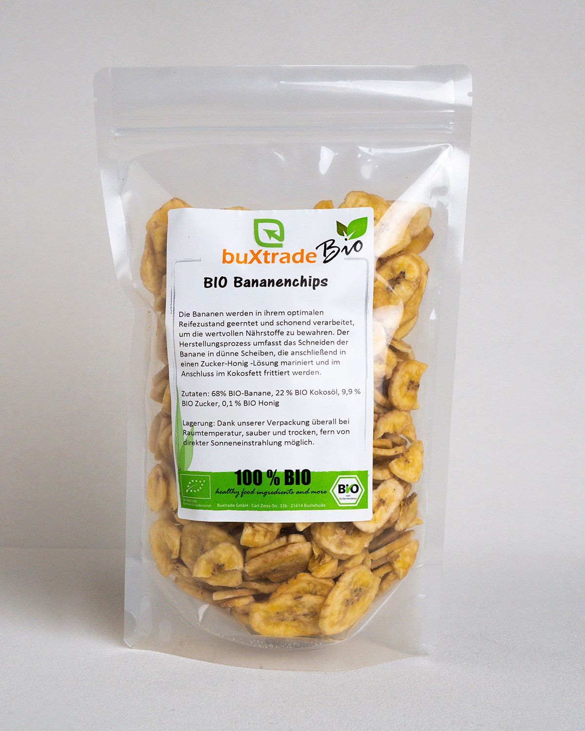 Transparente Tüte mit buXtrade Bio Bananenchips. Aufkleber mit Produktinformationen. Chips in der Tüte und auf einem weißen Podest.