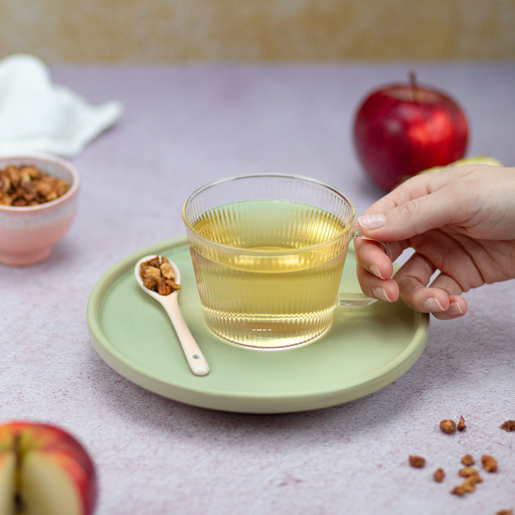 Glasbecher mit Tee auf Untertasse. Löffel mit Teestücken. Hand hält Becher. Äpfel im Hintergrund.
