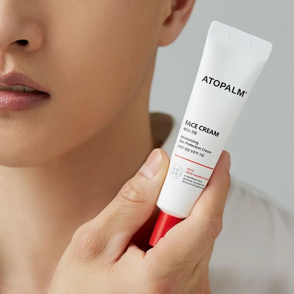 Person hält eine weiße Tube mit rotem Verschluss. Aufschrift ATOPALM und FACE CREAM. Hautpflegeprodukt.