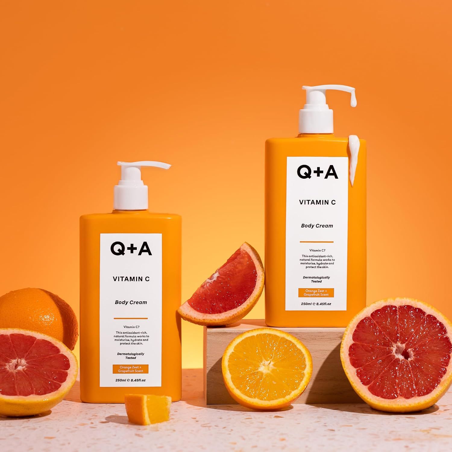 Q+A Vitamin C Body Cream Spenderflaschen, daneben Grapefruit und Orange.