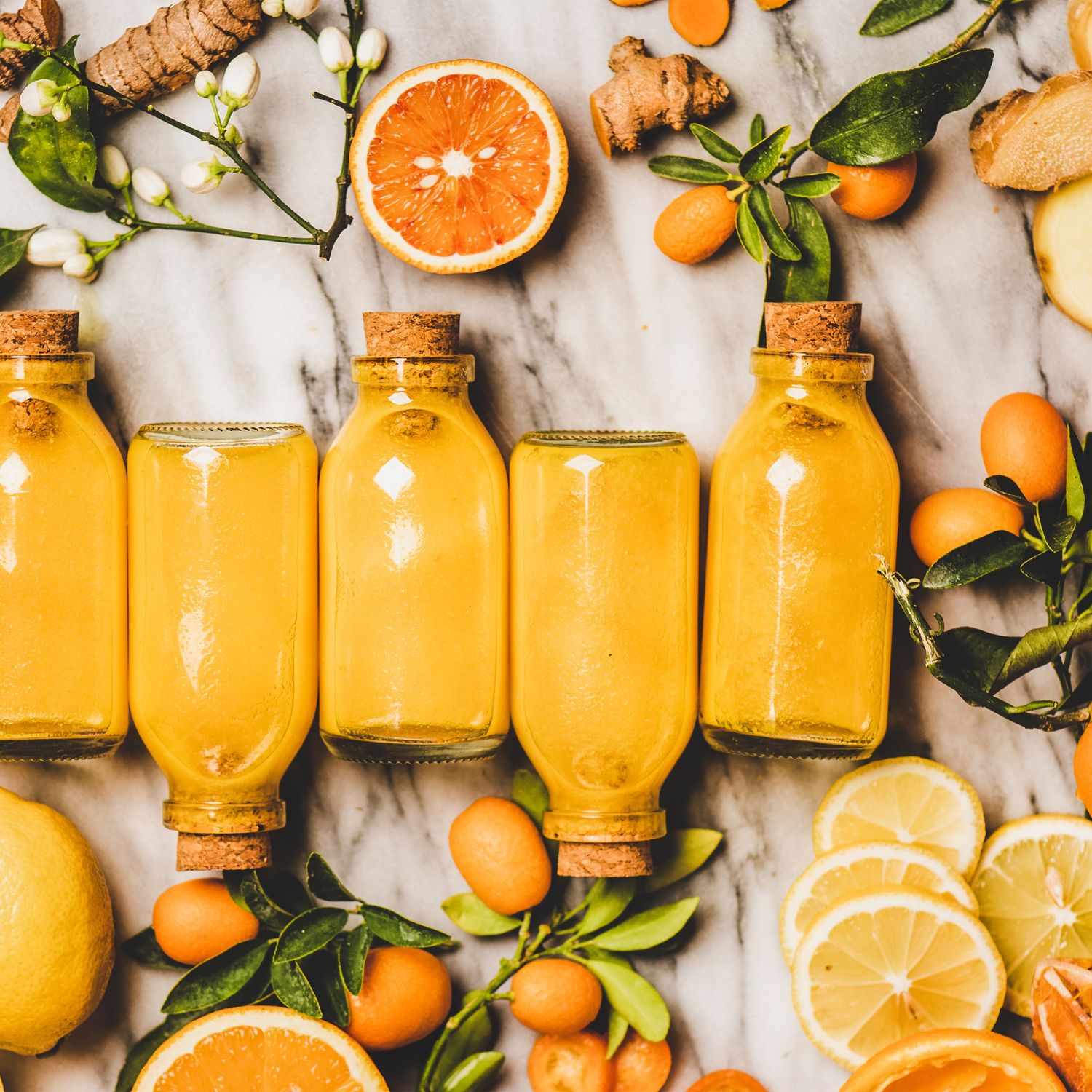 Reihe von Glasflaschen mit gelbem Inhalt, Korken. Zitrusfrüchte und Ingwerstücke umgeben die Flaschen.