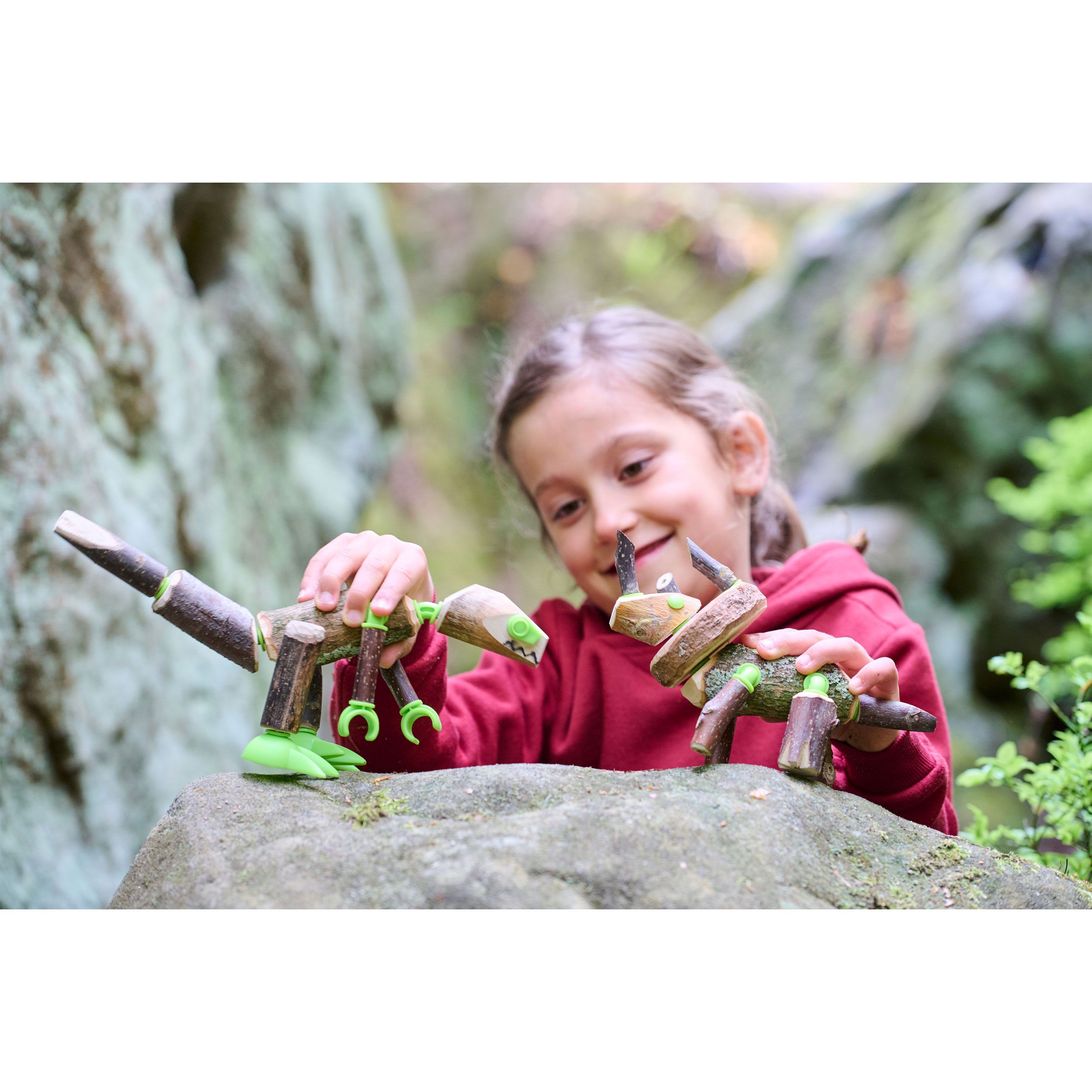 Ein Mädchen spielt mit zwei Dinosaurier-Konstruktionen aus Holz und grünen Verbindern. Sie sitzt auf einem Felsen im Freien.