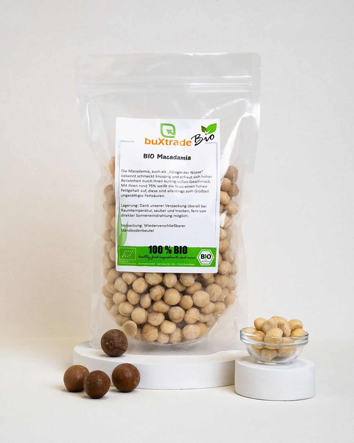 Transparente Verpackung mit buXtrade Bio Macadamia. Aufdruck: BIO, 100% BIO, Text in Deutsch. Zusätzlich Schale mit Nüssen.