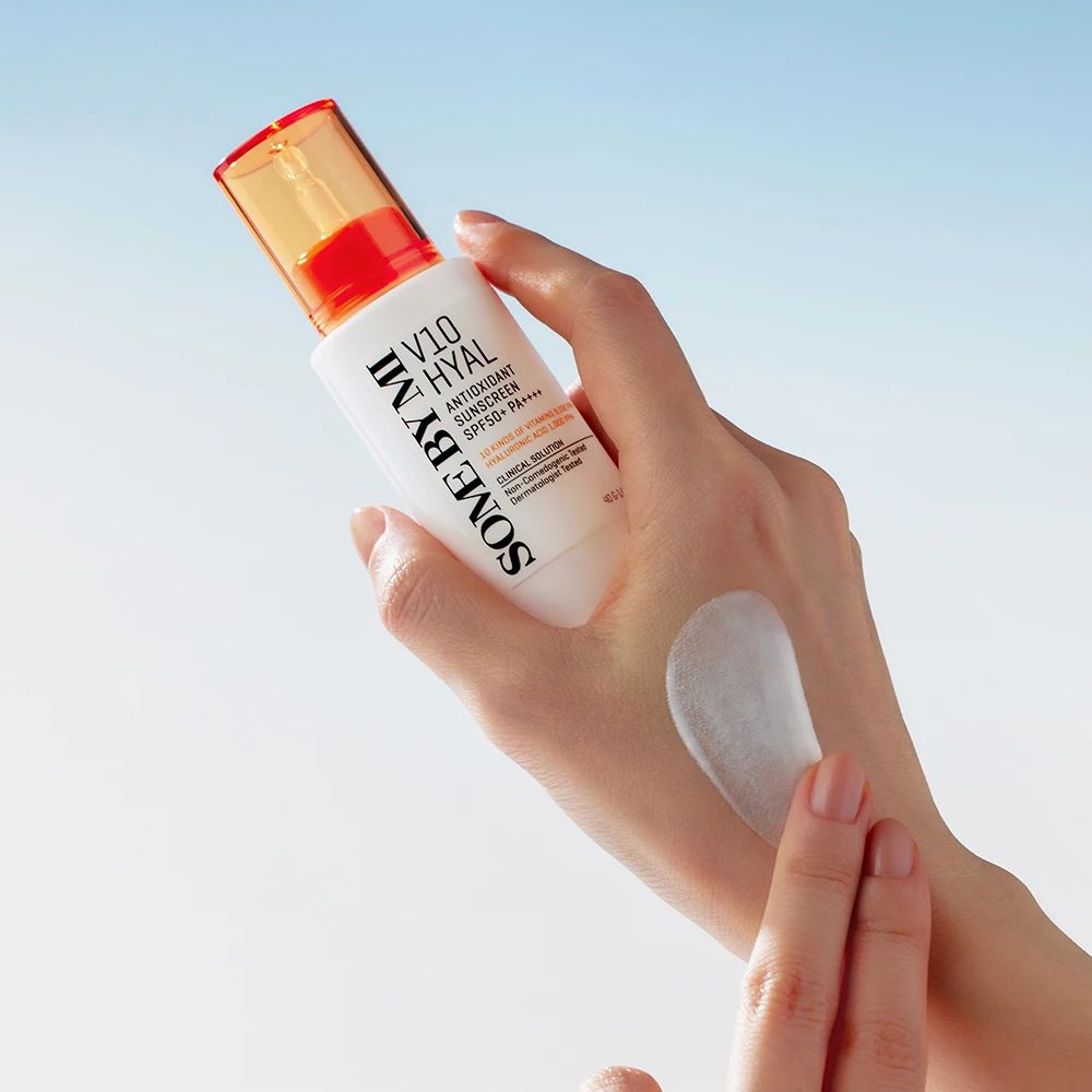 Weißer Flakon mit orangefarbenem Deckel. Produkt auf Hand aufgetragen. Text: SOME BY MI, V10 HYAL Antioxidant Sunscreen SPF50+.